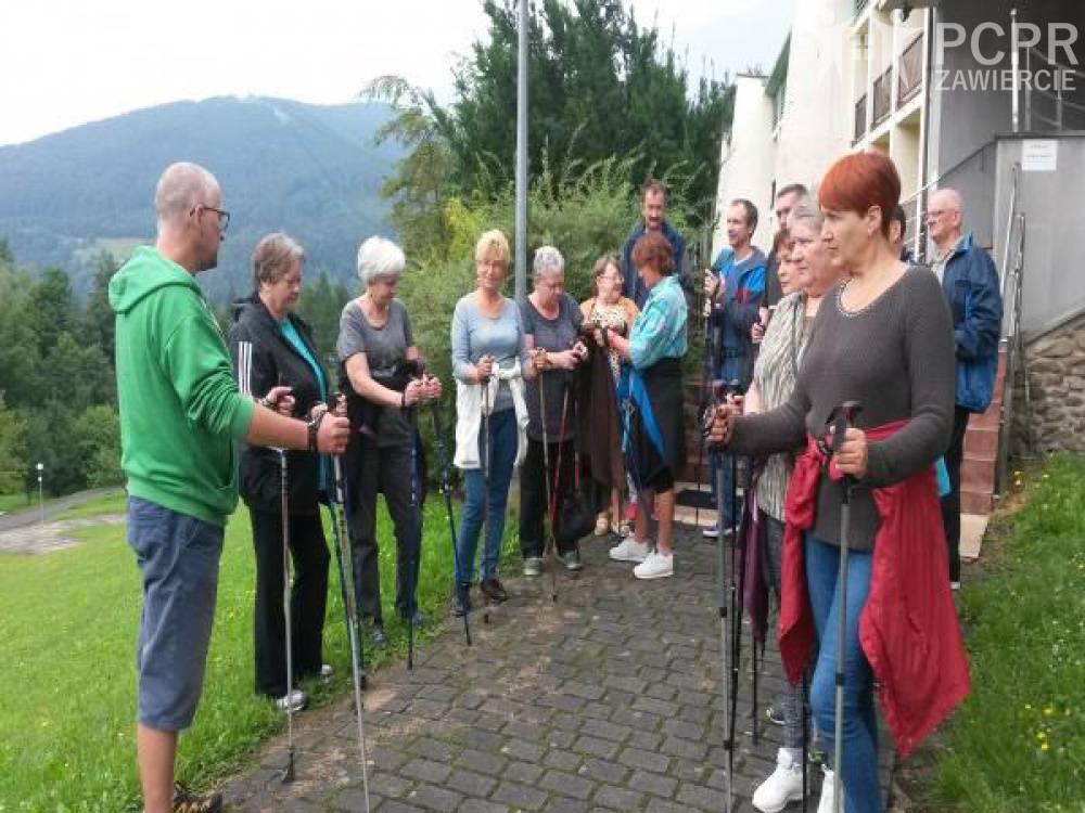 Zdjęcie: Uczestniczki i uczestnicy stoją z kijami do nordic walking i słuchają instruktora